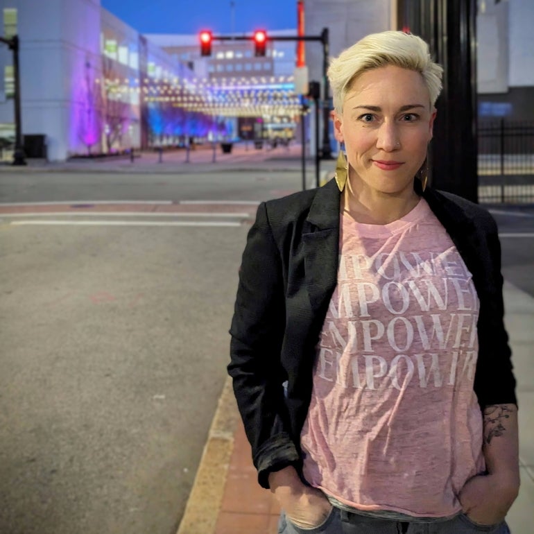 A woman wearing a black jacket and a pink shirt saying "Empower" stands in front of an urban intersection.