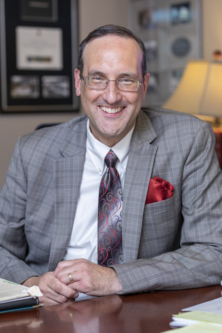 A man with glasses wearing a gray suit smiles at the camera.