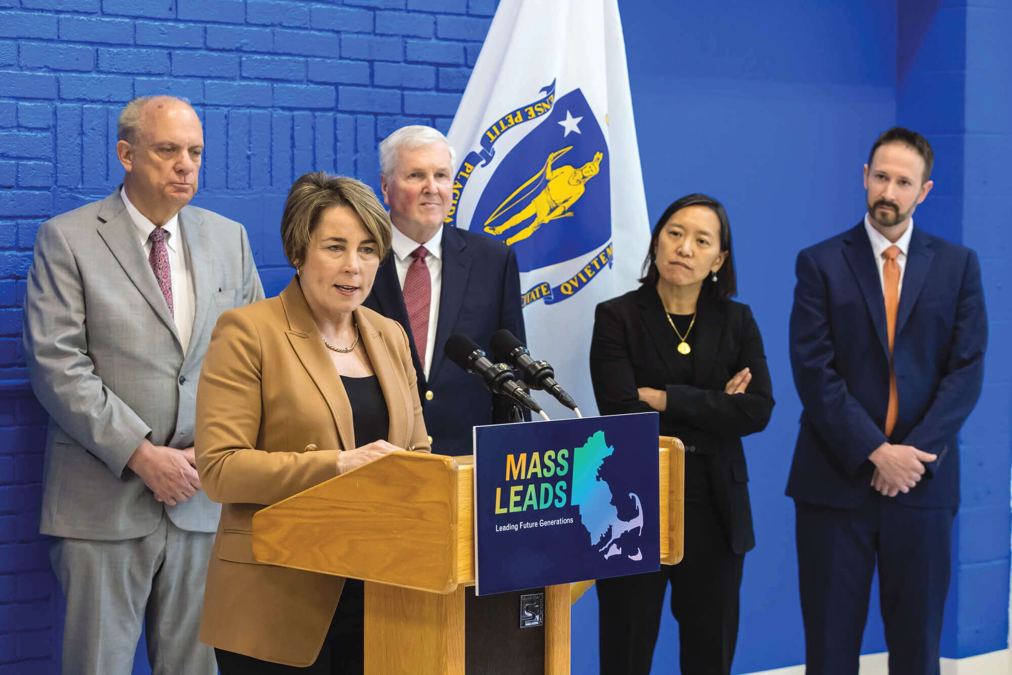 Five people stand behind a podium by a Massachusetts state flag