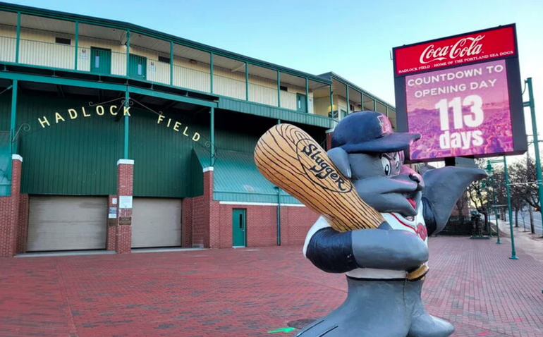 A statue of a seal holding a baseball bat stands in front of an electronic sign and baseball park