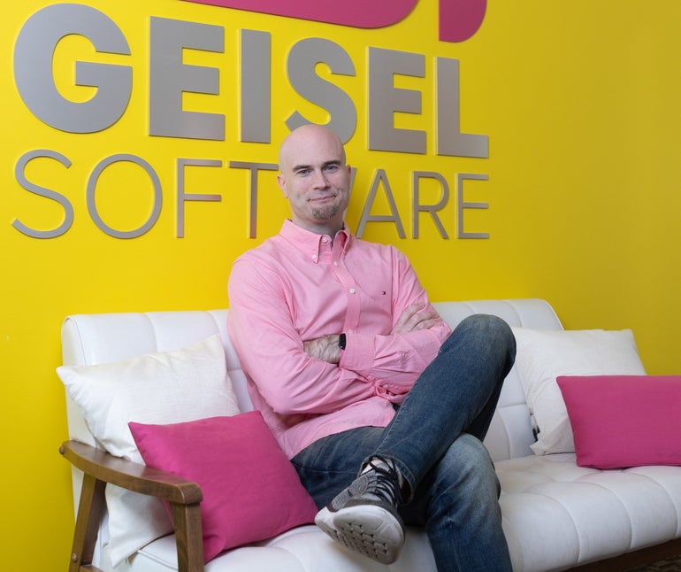 A man wearing a pink shirt and jeans sits on a white couch in front of a yellow wall with the Geisel Software logo on it.