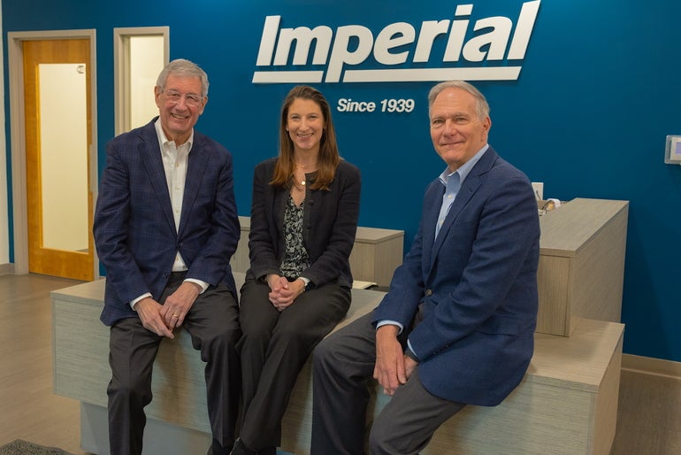 Two men in blue suits and a woman in a black suit sit in front of the Imperial Distributors logo.