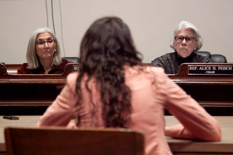 A woman sits at a desk in front of legislators