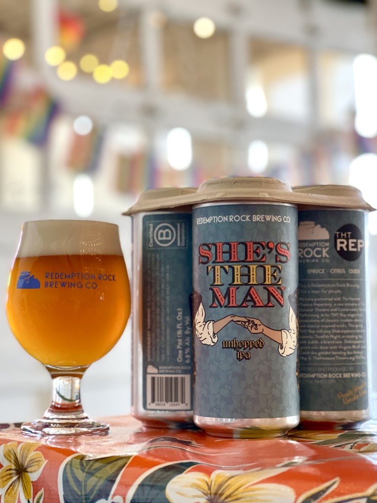 A glass of beer sits to the right of a group of three blue beer cans, the center can showing a cartoon image of hands being held underneath the beer's name "She's the Man" 