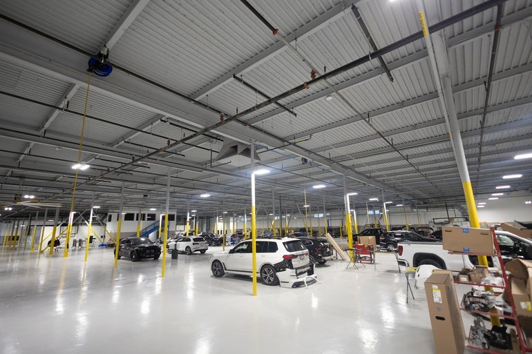 Inside a large warehouse with a white floor and yellow polls between cars to be repaired