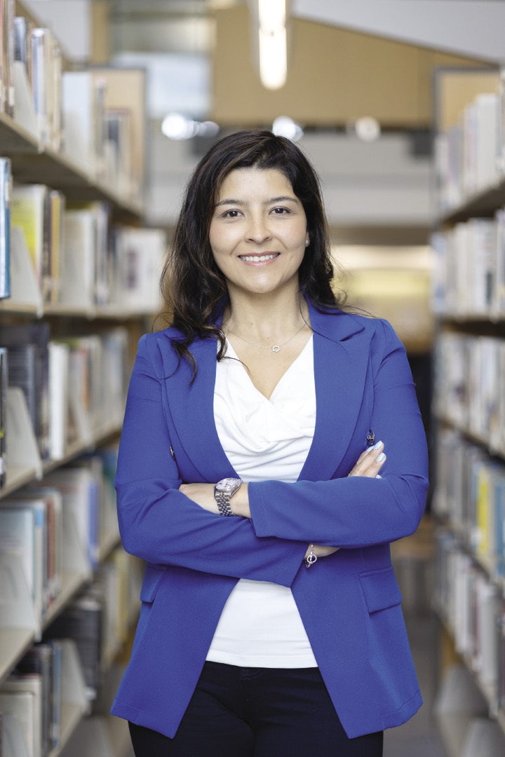A woman with dark brown hair wears a blue suit jacket and white blouse with folded arms and a silver watch in a library aisle.