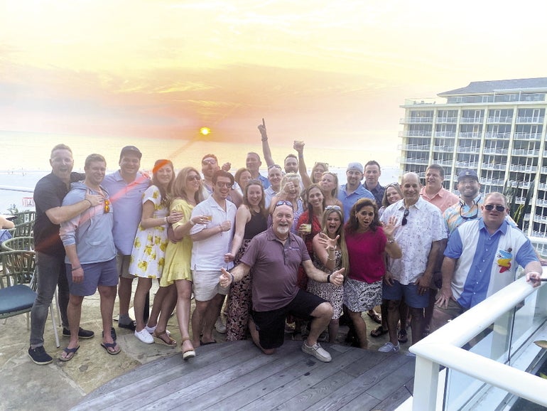 A large group of people stand in front of a setting sun at a resort.