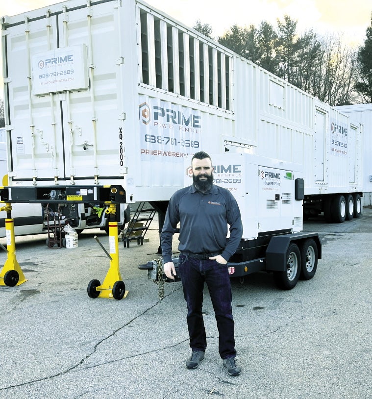 A man wearing jeans stands in front of a large generator.