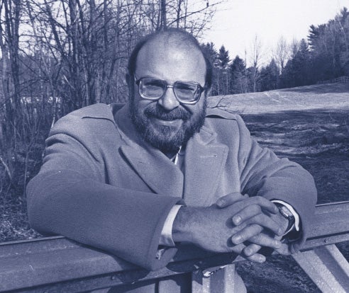 A man with glasses wearing an overcoat rests his folded hands on a railing.