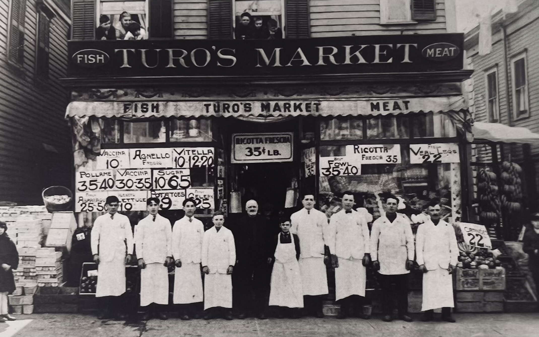 A black and white photo of men and boys in white jackets and aprons standing in front of a storefront with signs posted on the windows with prices of sold goods.