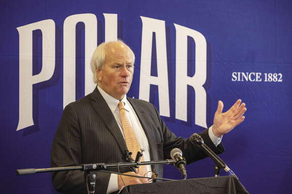 A man with white hair stands behind a podium with two microphones in front of a blue wall with "POLAR" written on the wall