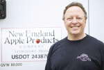 A man wears a black t-shirt standing in front of a sign reading "New England Apple Products"