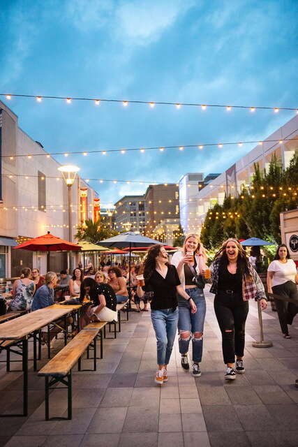 Three women walk towards the counter outside in a beer garden with string lights above them and a blue sky.