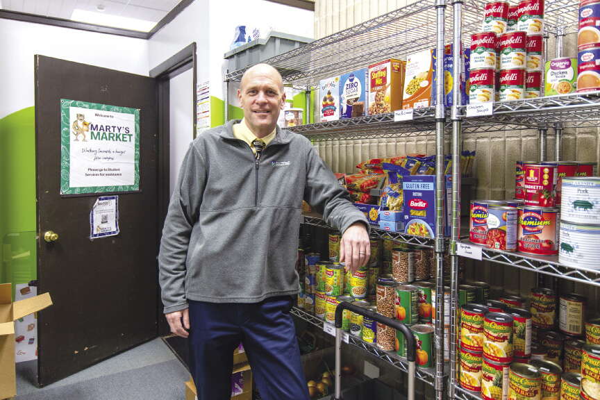 A mean wearing blue pants and a grey zip up sweater leans against metal shelving holding canned goods inside a food pantry.