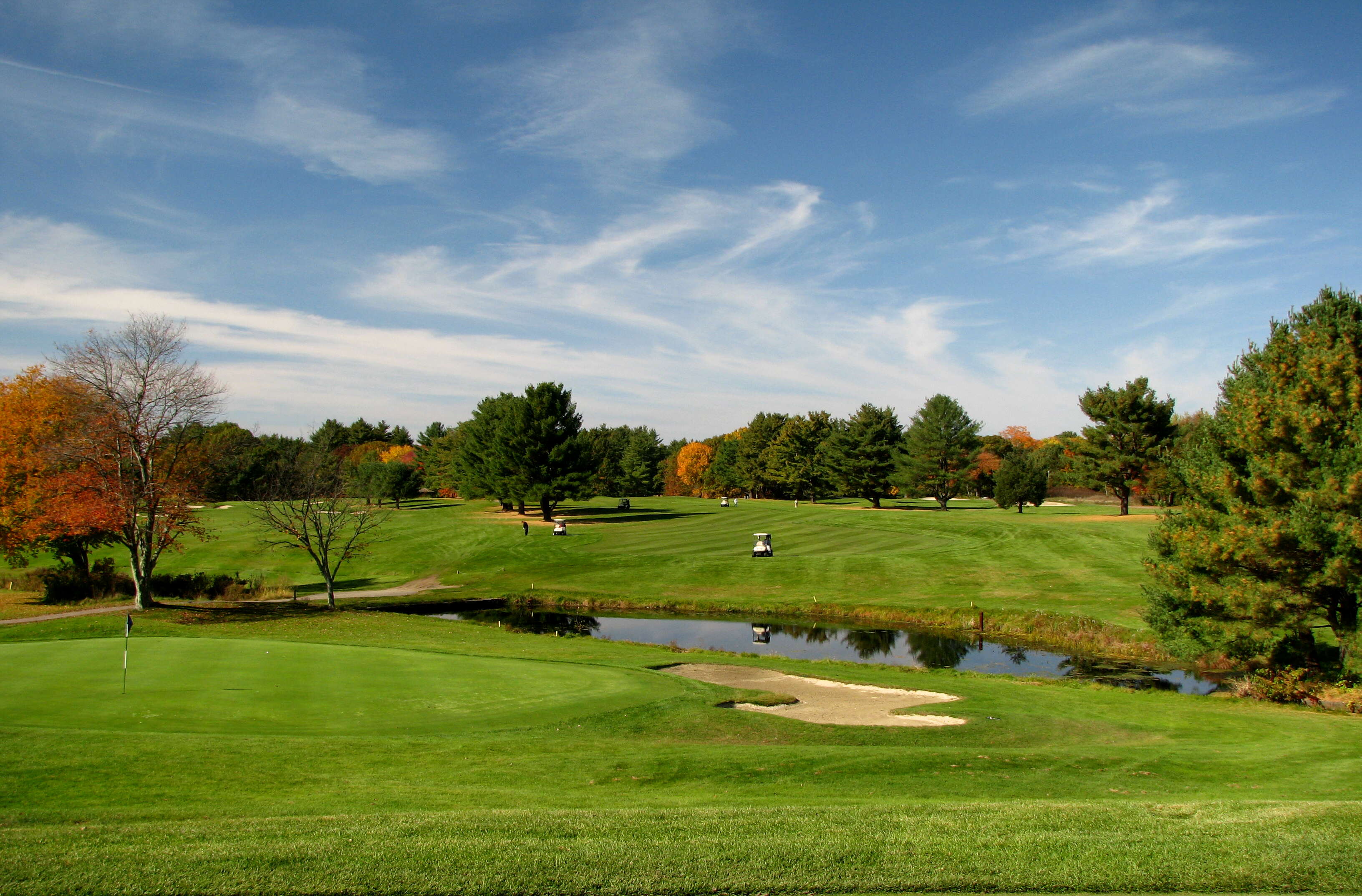 A green golf course with green and red trees throughout it under a blue and cloudy sky