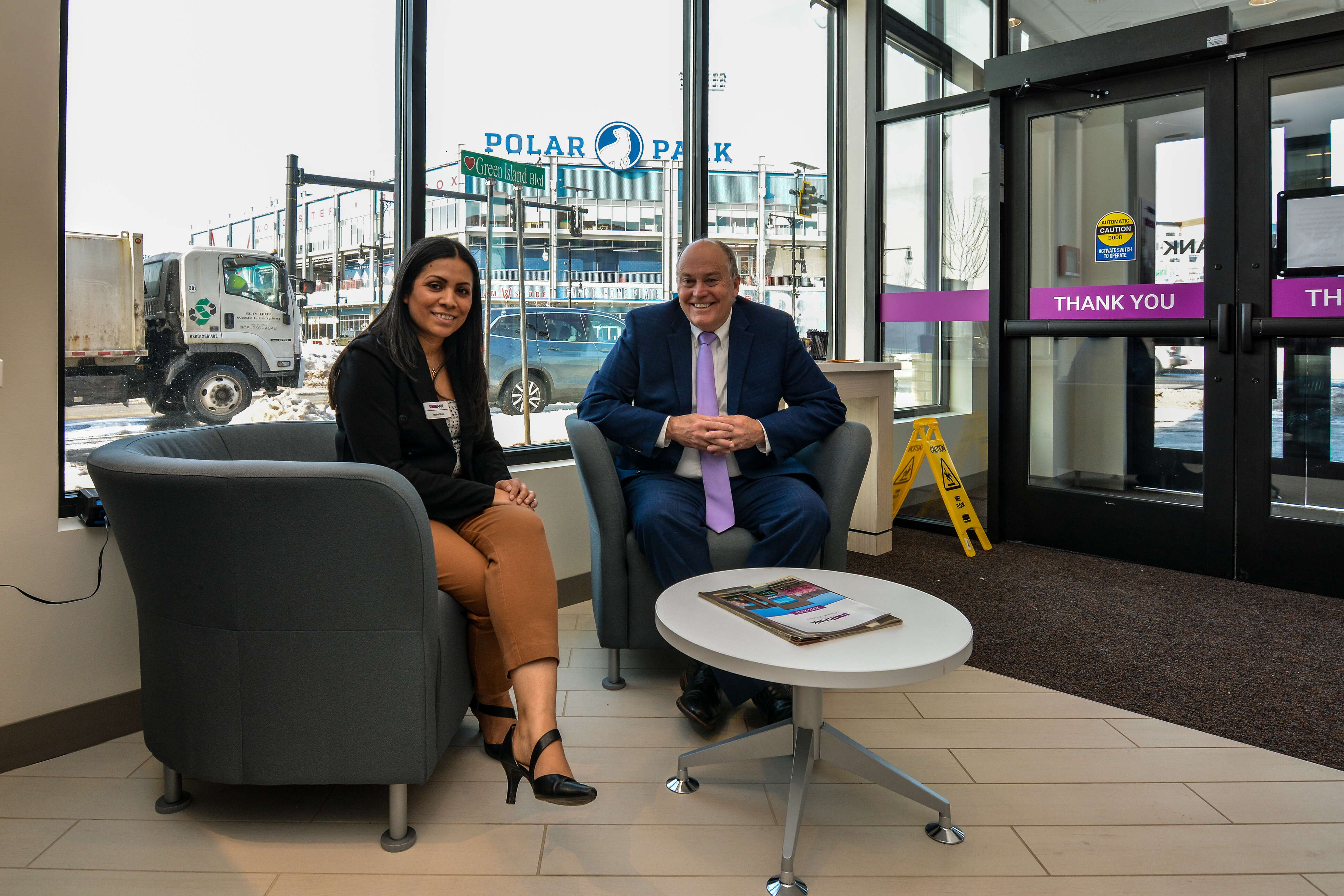 A woman with long dark hair wearing a black jacket and tan pants sits smiling at the camera at a round coffee table next to a man wearing a blue suit, a white button down, and a purple tie inside a bank smiling at the camera in front of a window opening to Polar Park baseball stadium.