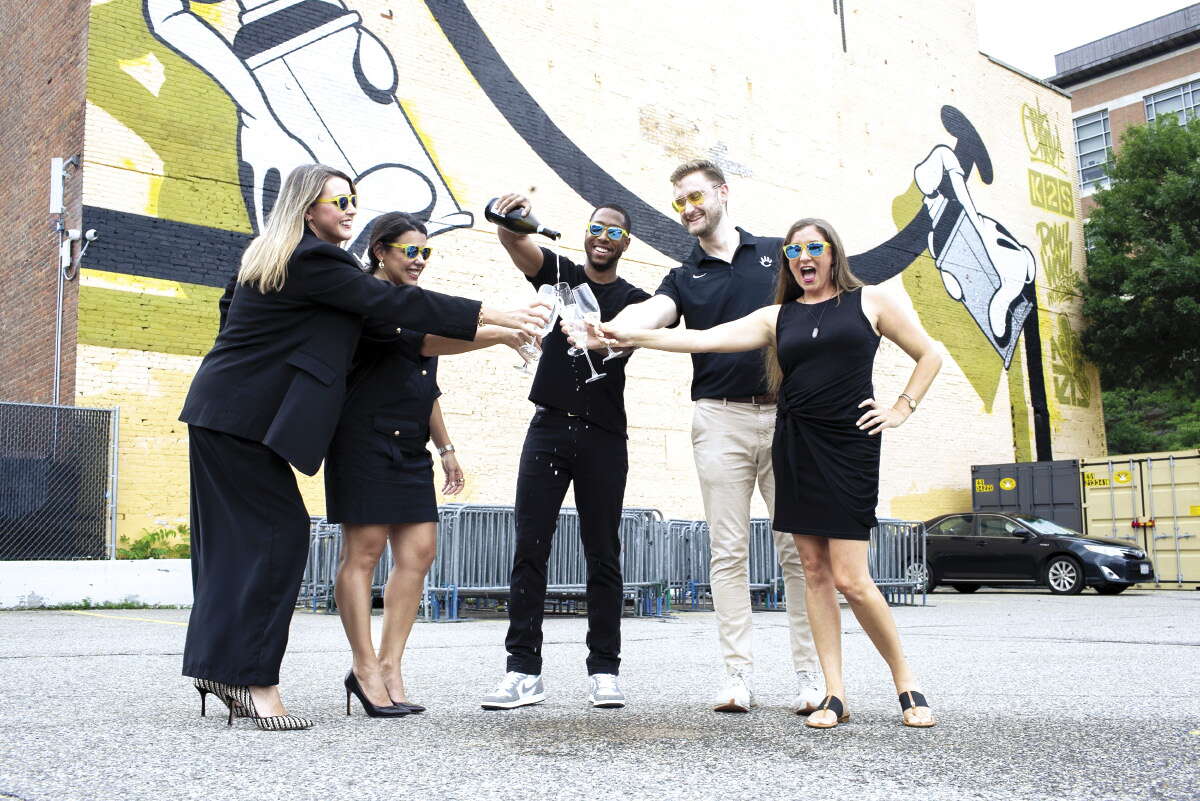 Three women and two men dressed in black pour champagne in front of a smiley face mural.