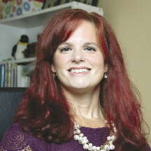 A woman with red hair wears a purple shirt and statement pearl necklace.