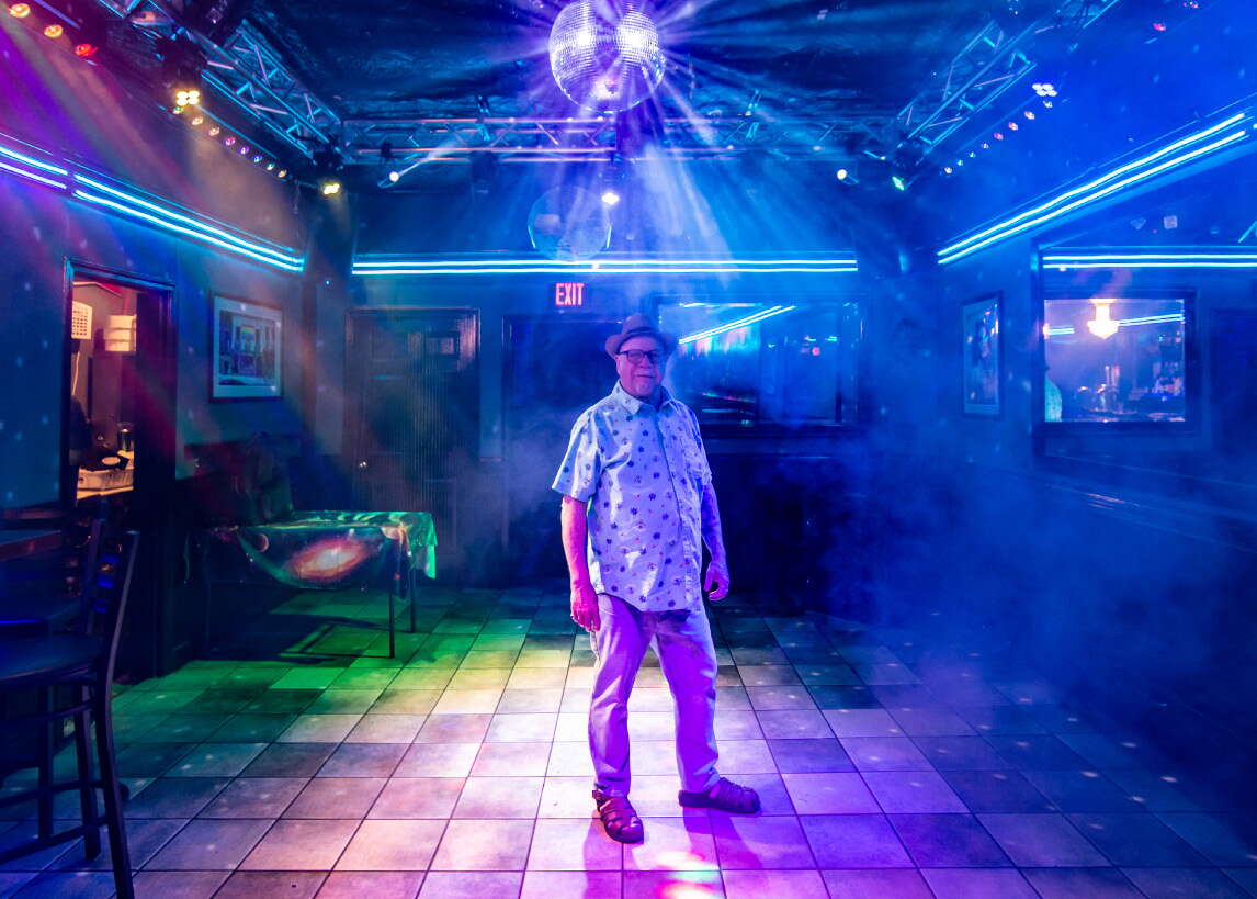 A man with a short sleeve button down and a fedora stand on a tiled dance floor amidst fog with lights shining in blue, green, and purple.