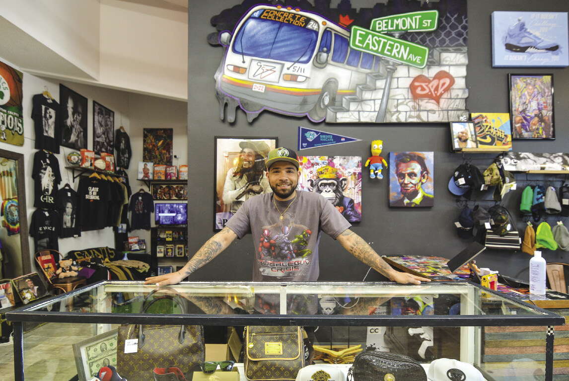 A man with a baseball cap, grey t-shirt, and tattoos on his arms stands with his hands on a glass case holding bags inside a store with art on the walls and racks of clothing and hats.