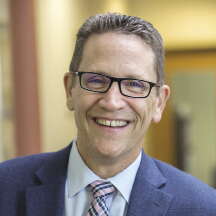 A man with glasses smiles for the camera in a dark blue suit jacket, light blue button down, and pink and blue patterned tie.