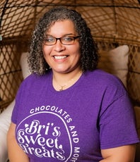 A woman with dark curly hair and glasses wears a bright purple t-shirt. 