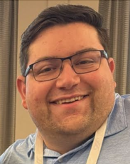 A man with short brown hair wears black glasses and a blue polo shirt.