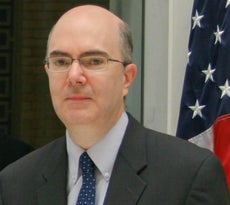 A man wears glasses, a grey suit jacket, light blue button down, and dark blue tie with white polka dots with an American flag behind him.