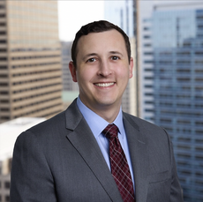 A man wears a dark grey suit jacket, a light blue button down, and a red plaid tie in front of a cityscape.