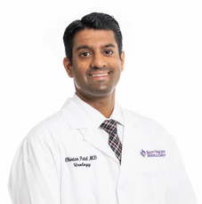 A man with gelled back dark hair wears a white lab coat and plaid tie.