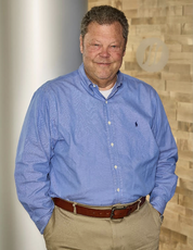 A man stands with his hands in his pockets wearing a light blue button down and khaki colored dress pants.