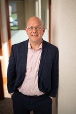 A man wears silver glasses, a pink checkered shirt, and a dark blue plaid suit jacket while leaning against a white wall.