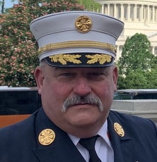 A man with a mustache wears a black and white hat with gold detailing.