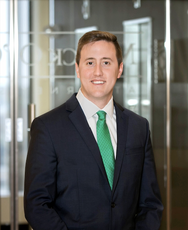 A man with light brown hair wears a dark blue suit jacket, white button down, and green tie.