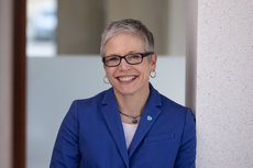 A woman with short white/grey hair wears black glasses, gold hoop earrings, and a blue blazer while leaning against a white wall.