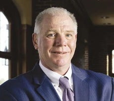 A man with white hair wears a dark blue suit jacket, white button down, and purple patterned tie.