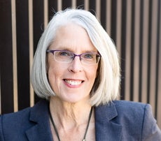 A woman with chin-length white hair smiles for the camera wearing purple glasses and a dark blue suit jacket.