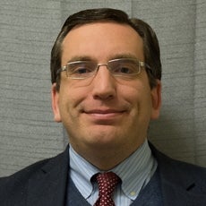 A man wears silver-rimmed glasses, a black suit jacket, dark blue vest, blue and white stripped button down, and red patterned tie.