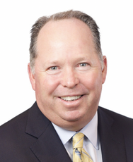 A headshot of a man with grey hair wears a black suit jacket, light blue button down, and yellow patterned tie