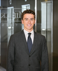 A man with dark brown hair wears a dark grey suit jacket, white button down, and blue tie.