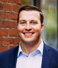 A man with light brown hair smiles for the camera in a dark blue suit jacket and blue and white checkered shirt.