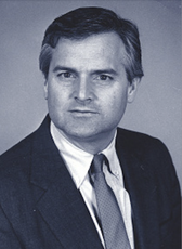 A black and white photo of a man wearing a dark suit jacket, white button down, and patterned tie