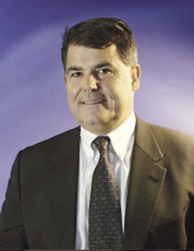 A man with short brown hair wears a black suit jacket, white button down, and a black patterned tie.