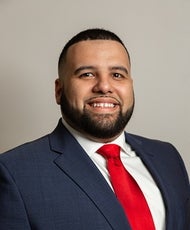 A man with a beard wears a blue suit with a red tie.