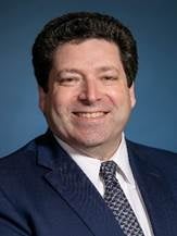 A man wears a dark blue suit jacket, white button down, and black and white tie.