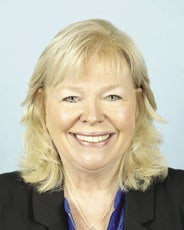 A woman with shoulder length layered blonde hair and bangs wears a black suit jacket and blue top.