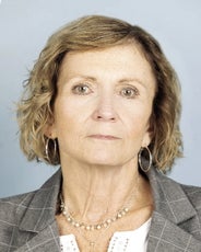 A woman with chin length blonde hair wears silver hoop earrings, a grey pinstripe suit jacket, and white top