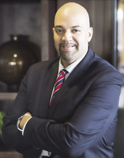 A hairless man stands with a goatee his arms folded wearing a black suit jacket, white button down, and red, white, and blue tie.