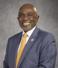 A bald man wears a blue suit jacket, white button down, and gold tie