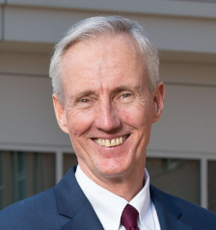 A headshot of a man with grey hair wearing a dark blue suit jacket, white button down, and red tie